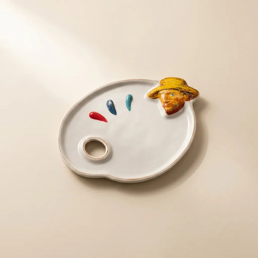 A white ceramic artist's palette-shaped tray with a small ring well, three paint-drip details in red, blue, and teal, and a sculpted figure of a painter wearing a yellow hat resting on the upper right edge, set against a warm beige background.