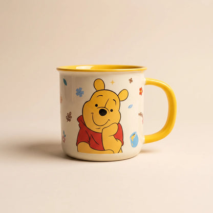 A cream-colored enamel mug featuring a cartoon illustration of Winnie the Pooh, with a bright yellow handle, yellow interior, and small scattered floral accents, set against a warm beige background.