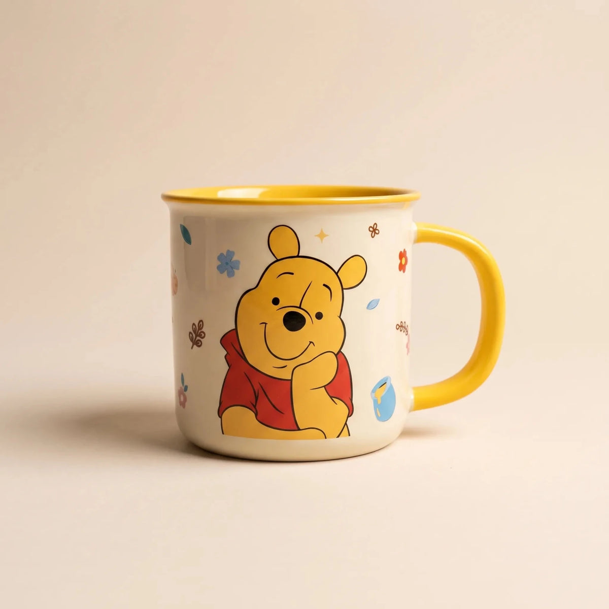 A cream-colored enamel mug featuring a cartoon illustration of Winnie the Pooh, with a bright yellow handle, yellow interior, and small scattered floral accents, set against a warm beige background.