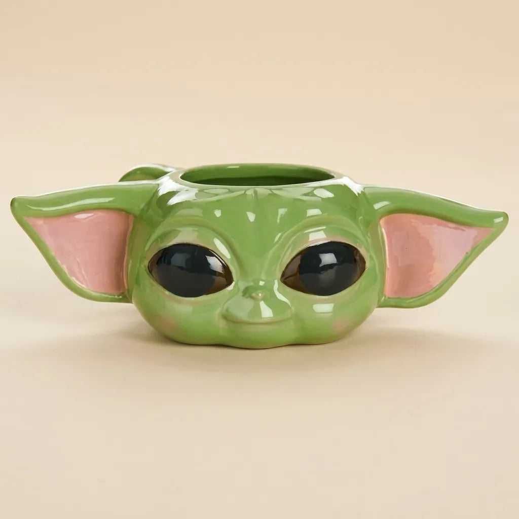 A green ceramic planter shaped like the character Grogu/Yoda's head, featuring pink inner ears and large black eyes, set against a solid beige background.